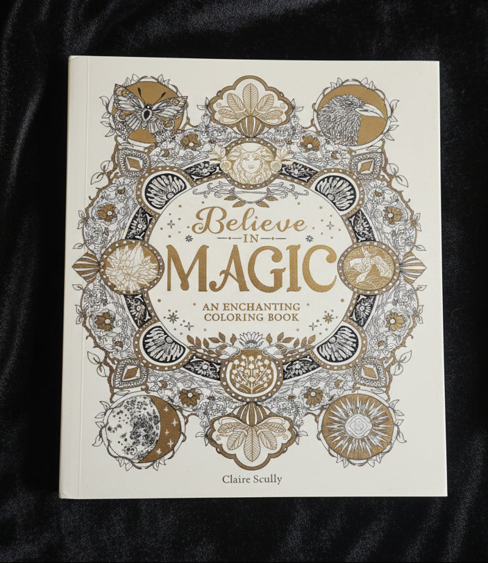 A hardcover coloring book titled 'Believe in MAGIC: An Enchanting Coloring Book' by Claire Scully, featuring intricate gold and gray line art illustrations on an off-white cover, held by a hand with red nail polish against a soft white fabric background.