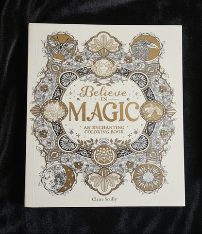 A hardcover coloring book titled 'Believe in MAGIC: An Enchanting Coloring Book' by Claire Scully, featuring intricate gold and gray line art illustrations on an off-white cover, held by a hand with red nail polish against a soft white fabric background.