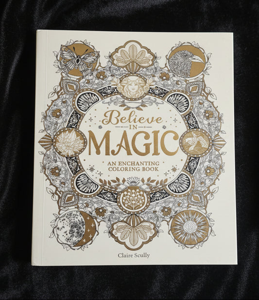 A hardcover coloring book titled 'Believe in MAGIC: An Enchanting Coloring Book' by Claire Scully, featuring intricate gold and gray line art illustrations on an off-white cover, held by a hand with red nail polish against a soft white fabric background.