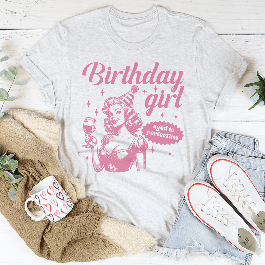 White t-shirt with pink 'Birthday girl' design on a white surface with casual items.
