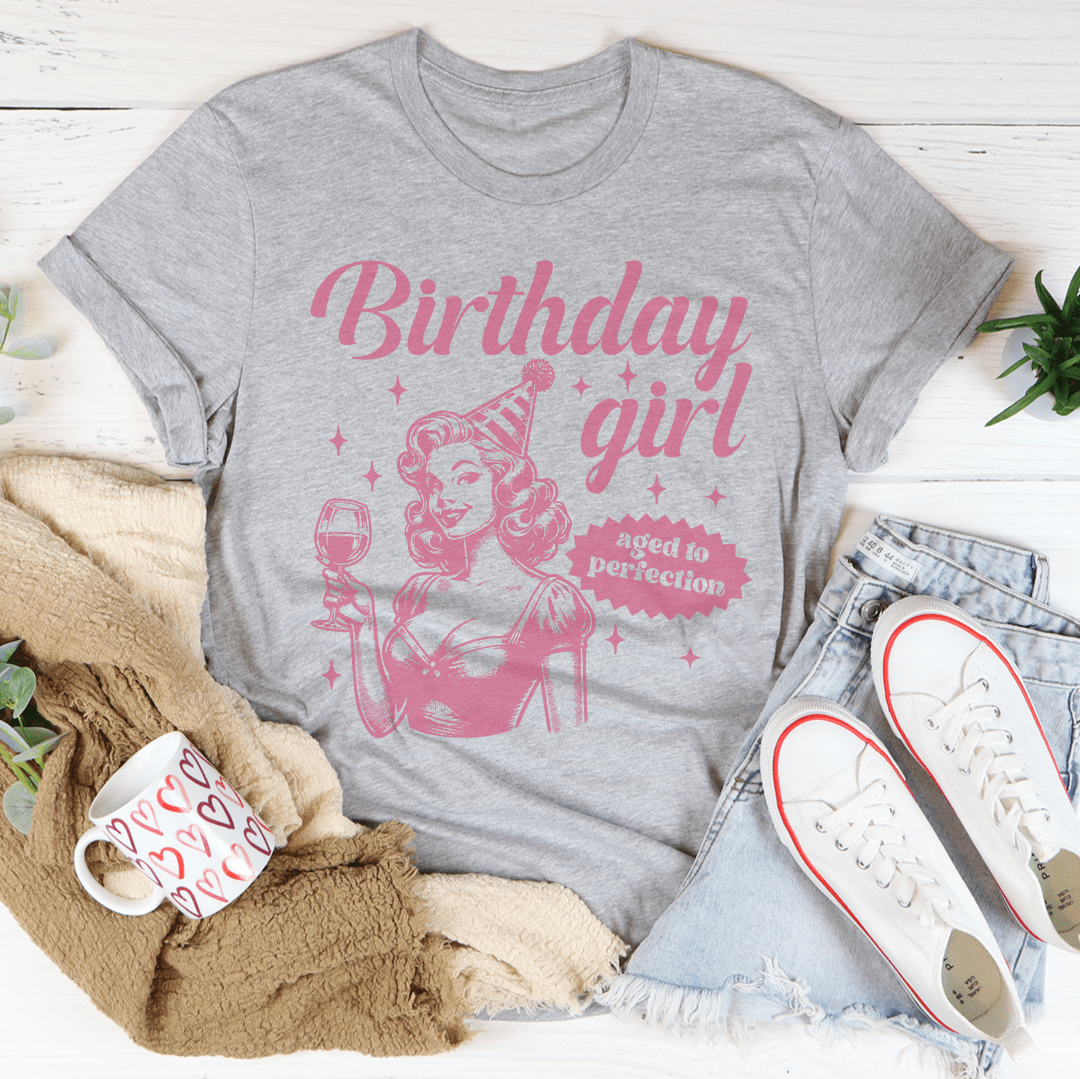 Gray t-shirt with pink 'Birthday girl' design on a white surface with accessories.