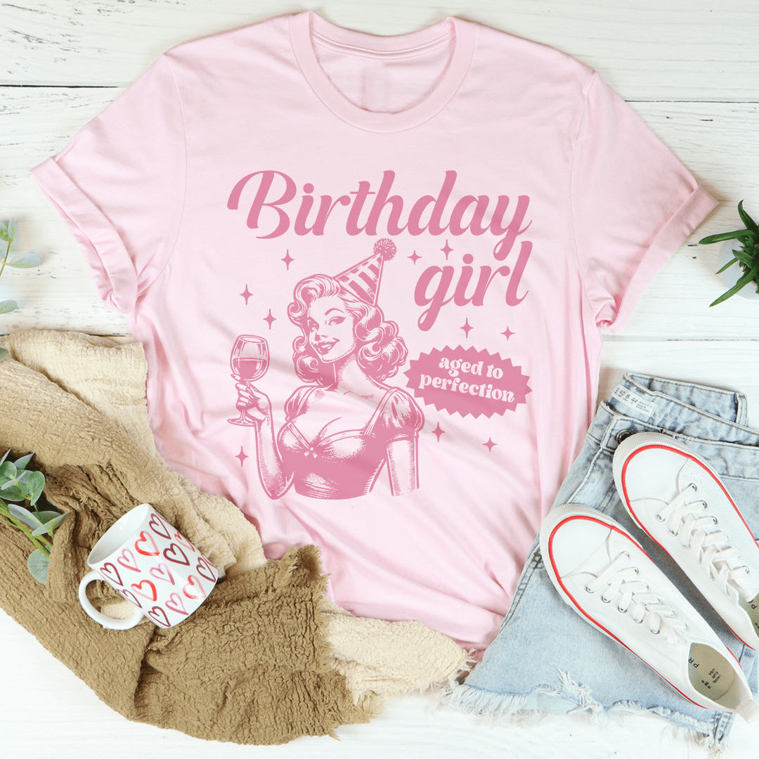 Pink t-shirt with 'Birthday girl aged to perfection' design on a white surface with accessories.