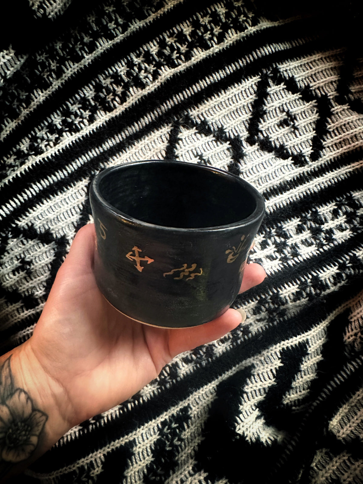 Black cup with gold designs held by a hand against a patterned fabric background
