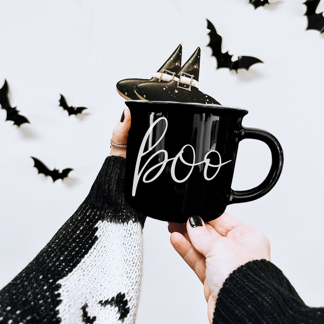 Black mug with 'boo' text held by a person, with decorative bats in the background