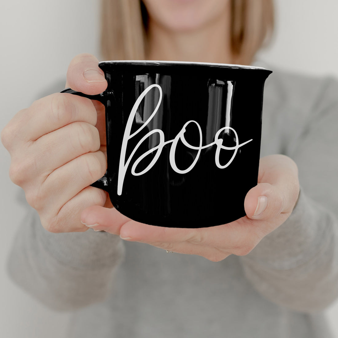 Person holding a black mug with 'boo' written on it against a neutral background