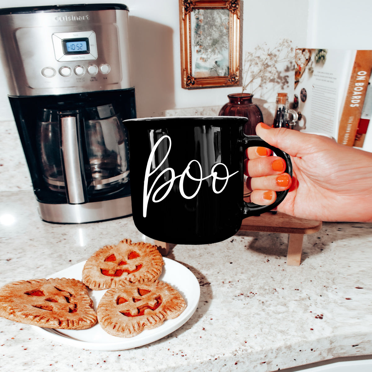 Person holding a black mug with 'boo' on it next to pumpkin pies and a coffee maker.
