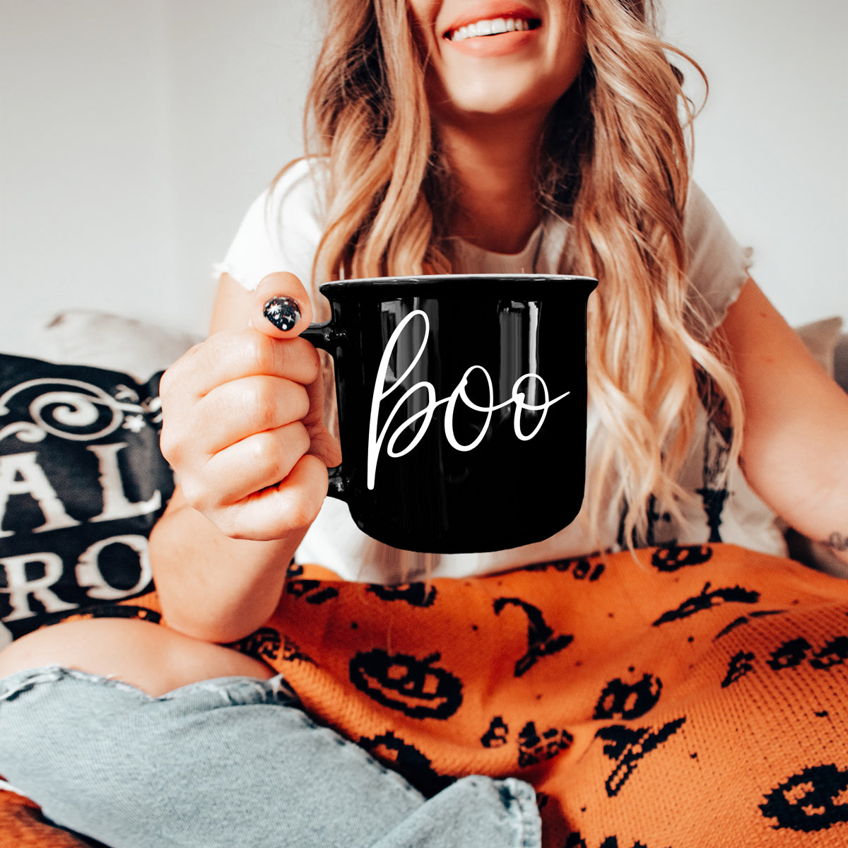 Person holding a black mug with 'boo' on it, wearing an orange Halloween-themed blanket.