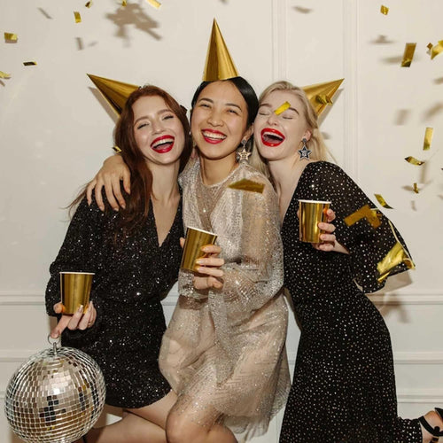 Three women celebrating with gold party hats and drinks, surrounded by confetti.