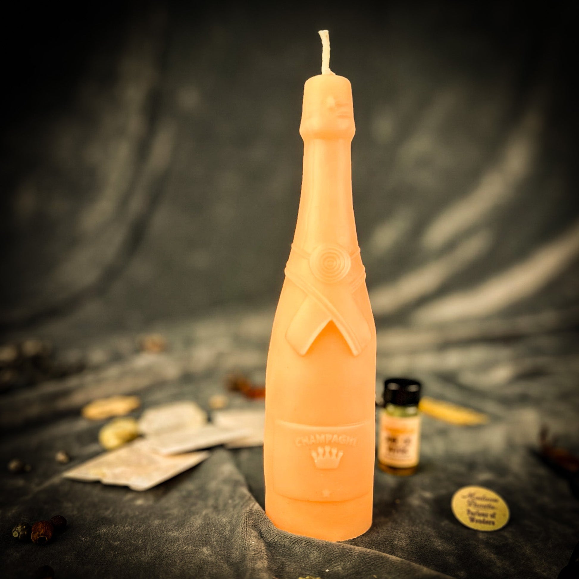 A tall, sculptural candle shaped like a champagne bottle stands upright on a dark draped surface. The candle is a pale peachy orange color with a small white wick.