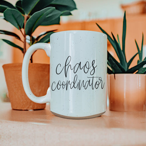 White mug with 'chaos coordinator' text in a home setting with plants