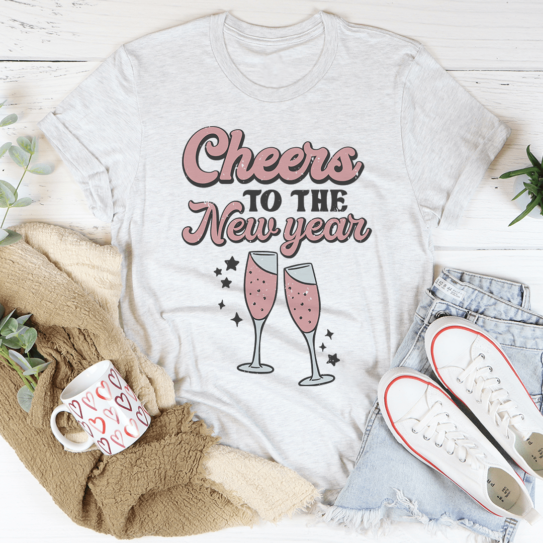 White t-shirt with 'Cheers to the New Year' text and champagne glass graphics, surrounded by casual items on a light wooden surface.