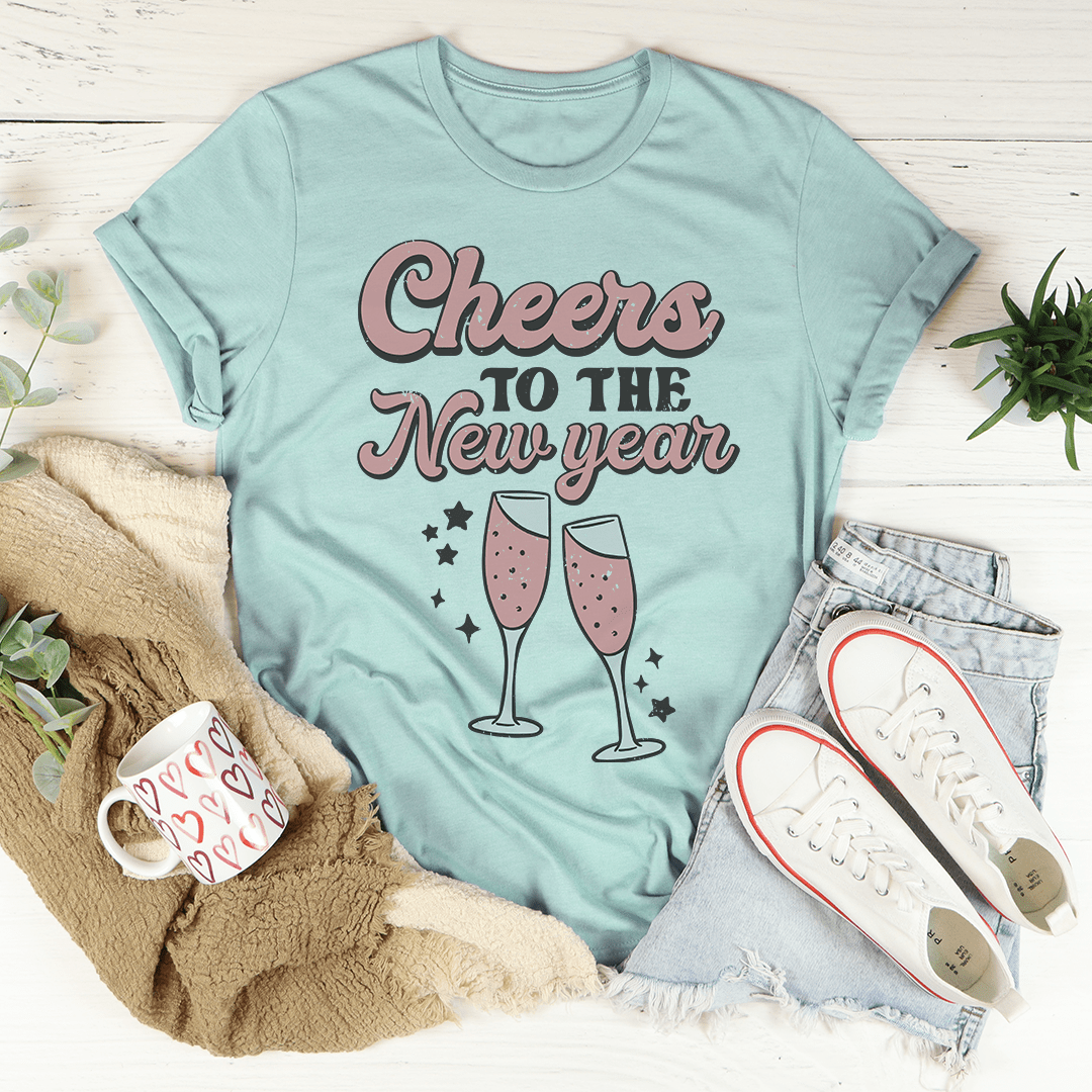 Light blue t-shirt with 'Cheers to the New Year' text and champagne glass graphics, surrounded by a mug, scarf, and shoes on a light wooden surface.