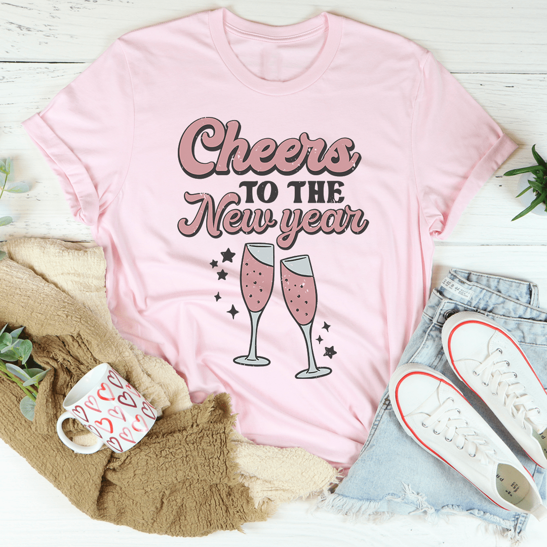 Pink t-shirt with 'Cheers to the New Year' text and champagne glass graphics, surrounded by casual clothing and decor.