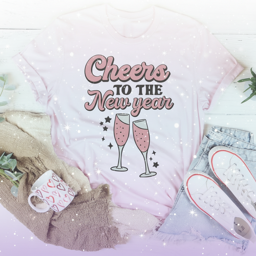 Pink t-shirt with 'Cheers to the New Year' text and champagne glass graphics, surrounded by casual clothing and decor.