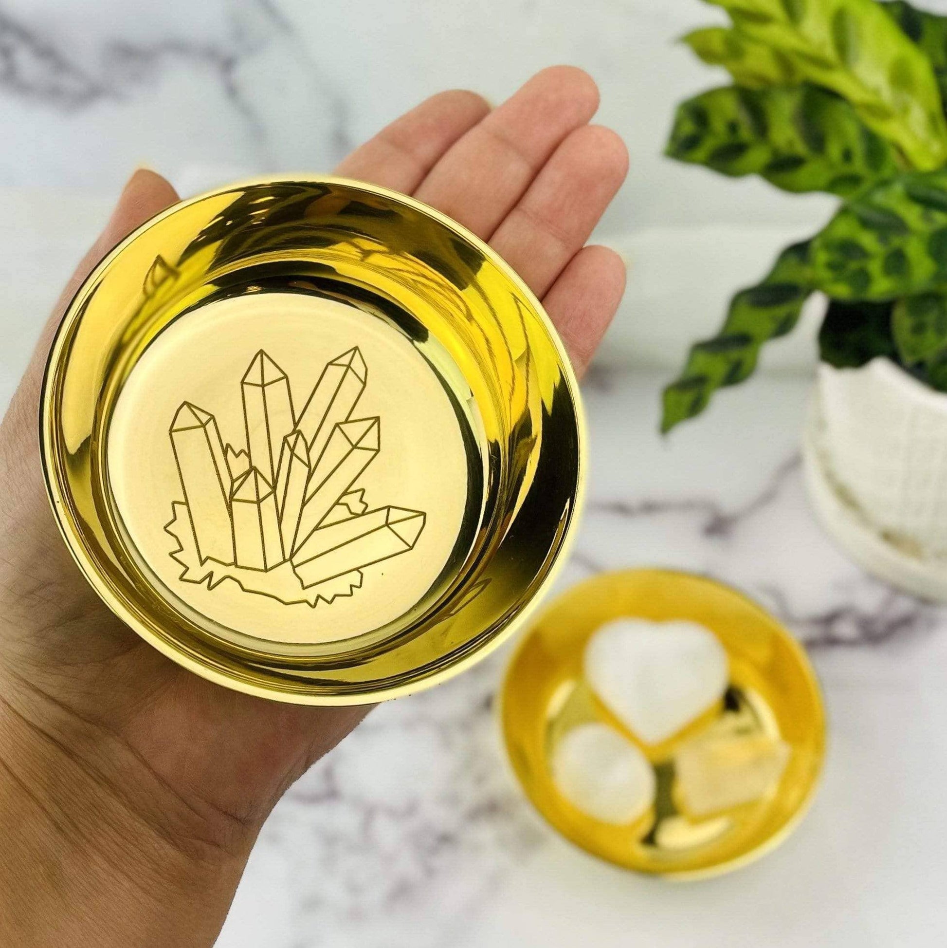 Gold dish with crystal design held by a hand on a marble surface with plants.