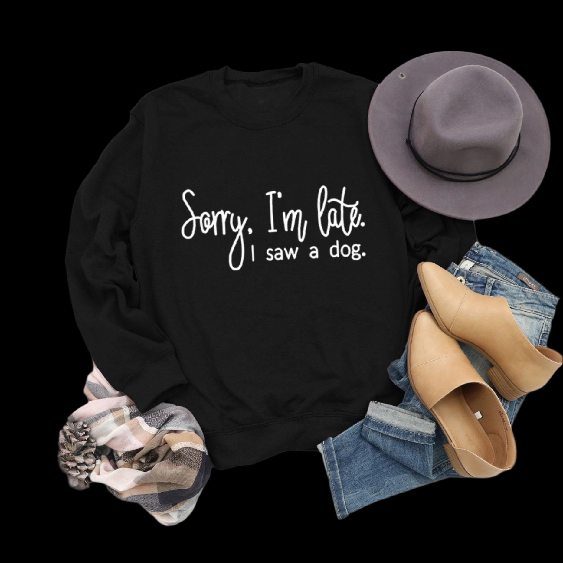 Black sweatshirt with 'Sorry, I'm late. I saw a dog.' text, paired with jeans, boots, and a hat on a wooden surface.