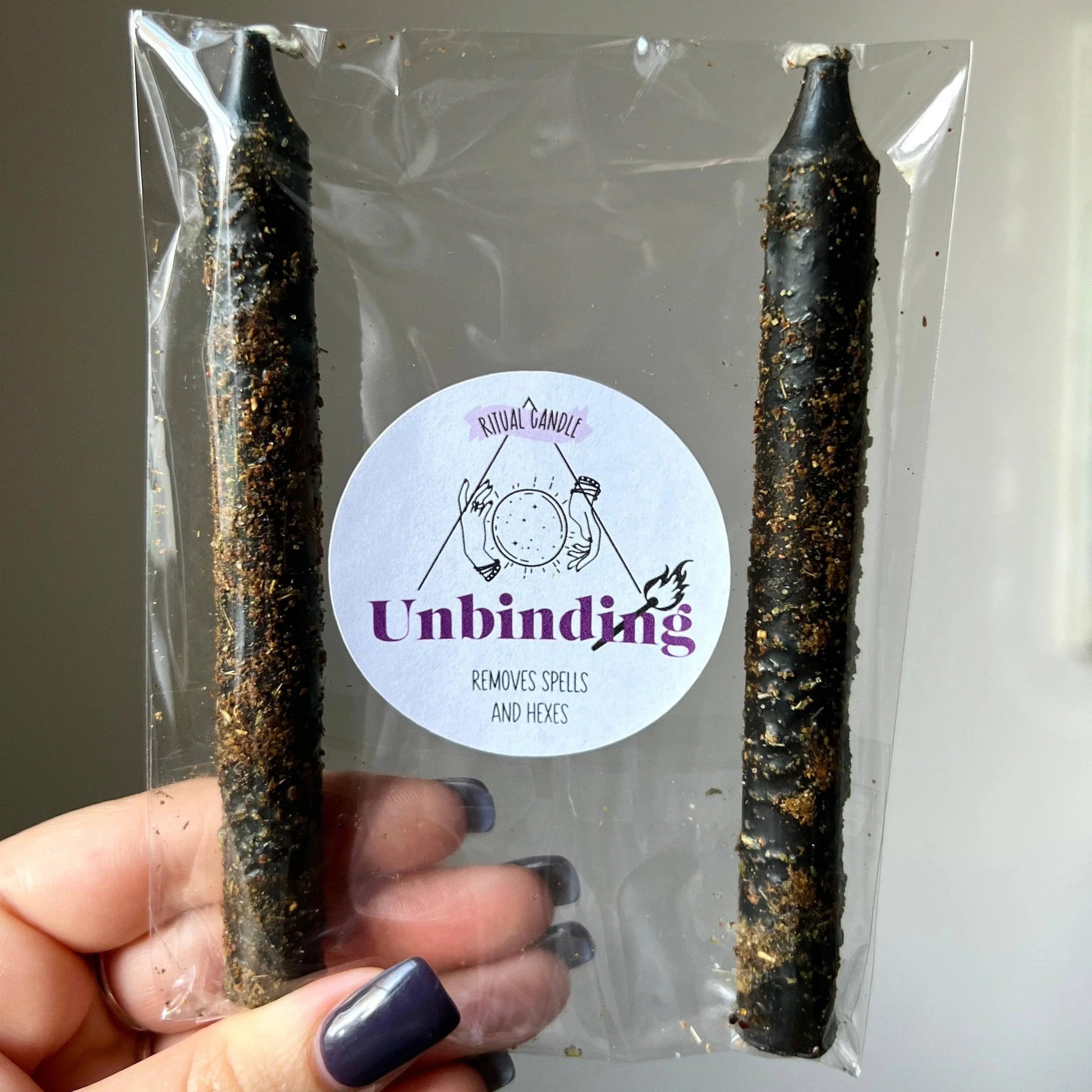 Two black candles in clear packaging with a 'Unbinding' label held by a hand.