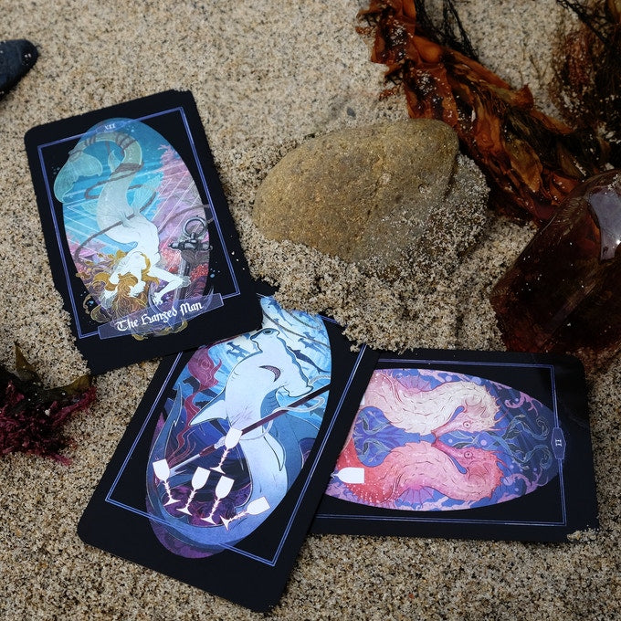 Three illustrated oracle or tarot-style cards are arranged on sand with small natural props, including a rock, dried leaves, and a crystal. The cards have dark backs with colorful fantasy artwork in blue, pink, purple, and cream tones.