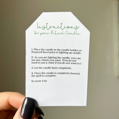 Hand holding a white card with green text instructions for a ritual candle against a neutral background