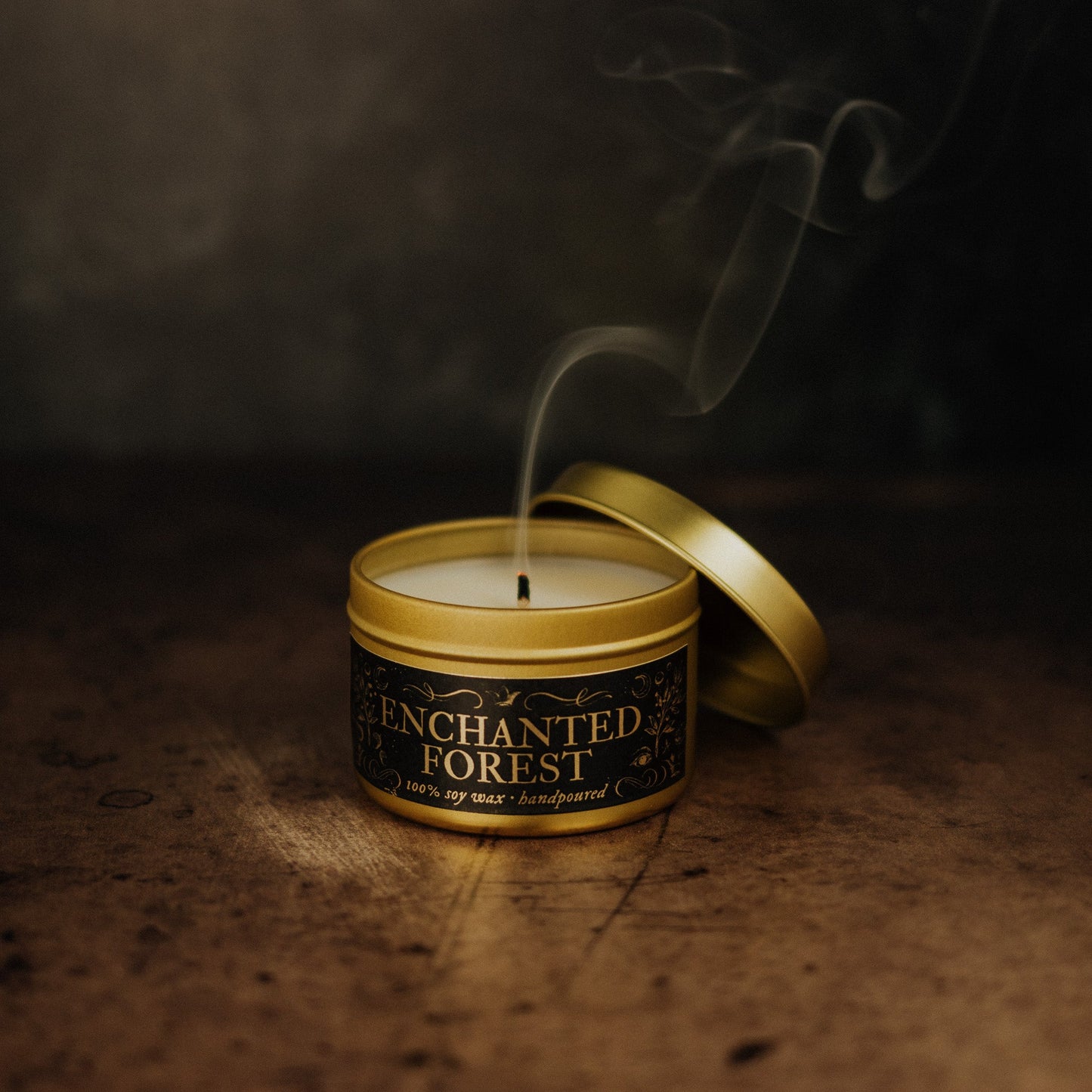 Candle in a tin labeled 'Enchanted Forest' with smoke against a dark background