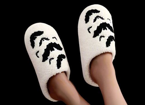White slippers with black paw prints worn by a person sitting on a pink cushion.
