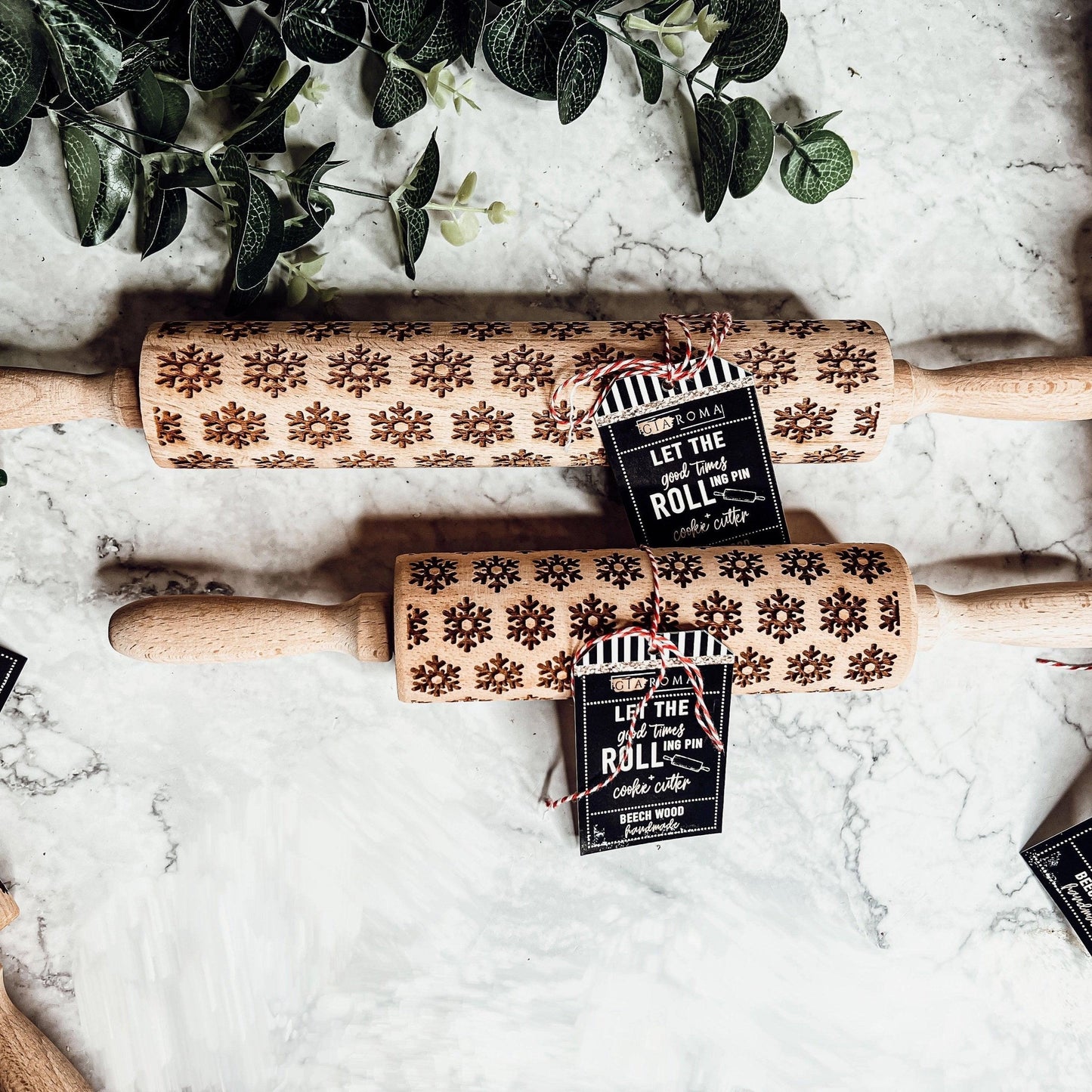 Two wooden rolling pins with snowflake patterns on a marble surface, accompanied by greenery.