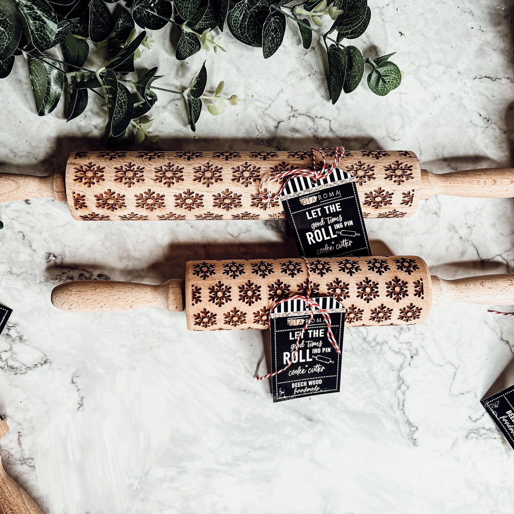 Two wooden rolling pins with snowflake patterns on a marble surface, accompanied by greenery.