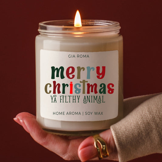 Candle with a 'Merry Christmas Ya Filthy Animal' label held by a hand against a dark background