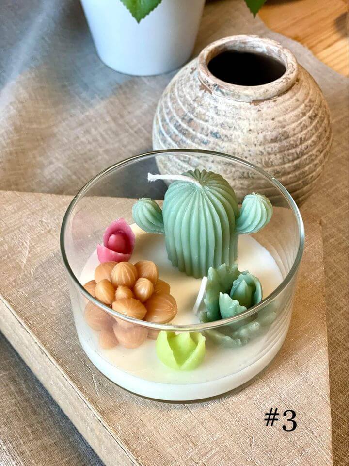 Candle with cactus-shaped wax figures in a glass container on a wooden surface.