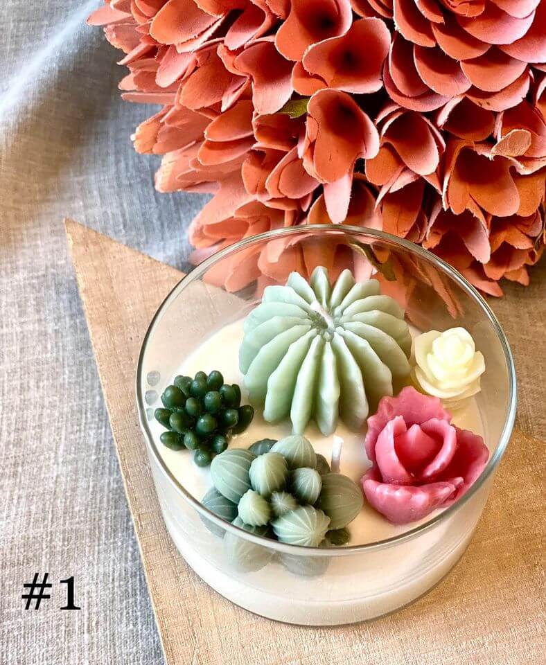 Decorative candle with colorful succulent-like sculptures in a glass container, with pink flowers in the background.