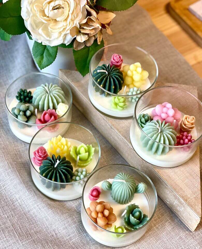 Colorful succulent-shaped candles in glass holders on a textured surface with flowers in the background.