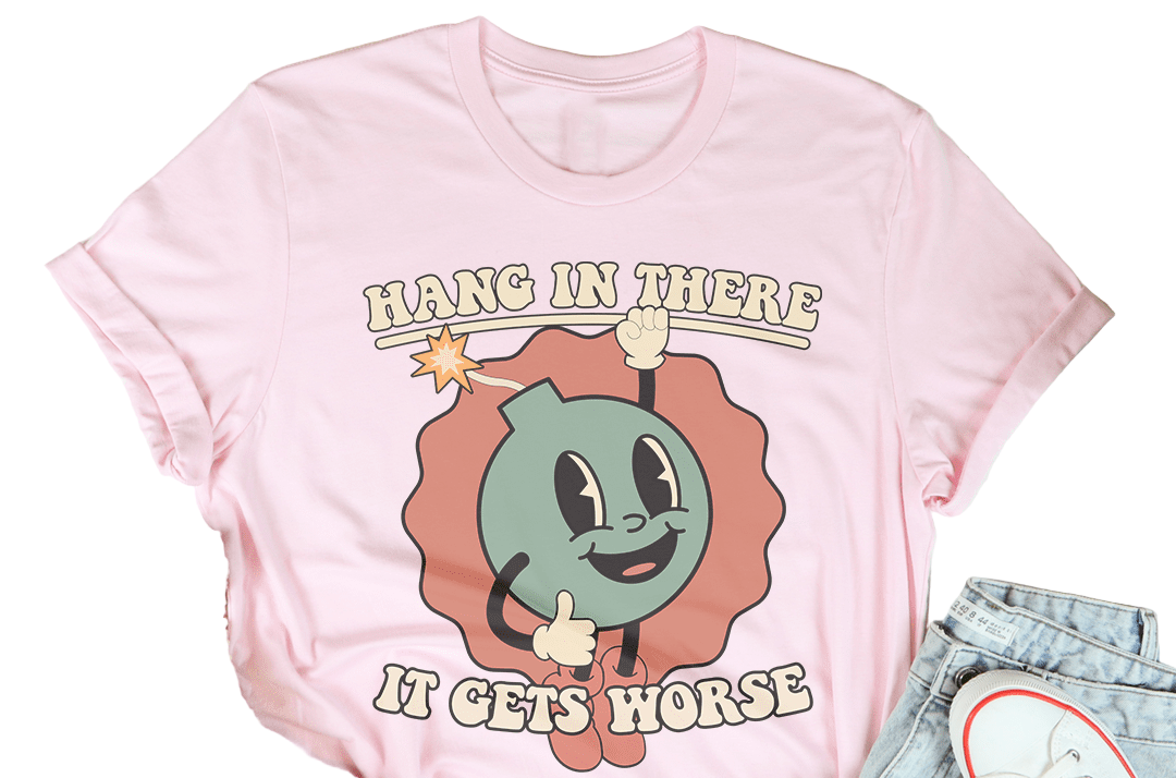 Pink t-shirt with cartoon character and text 'Hang in there, it gets horse' on a wooden surface with casual items.