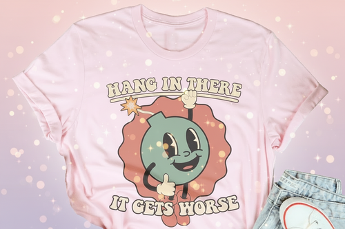 Pink t-shirt with cartoon character and text 'Hang in there, it gets horse' on a wooden surface with casual items.