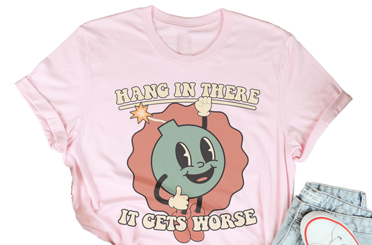 Pink t-shirt with cartoon character and text 'Hang in there, it gets horse' on a wooden surface with casual items.