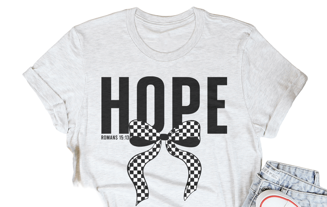 White t-shirt with 'HOPE' and checkered bow design, placed on a wooden surface with casual items around.