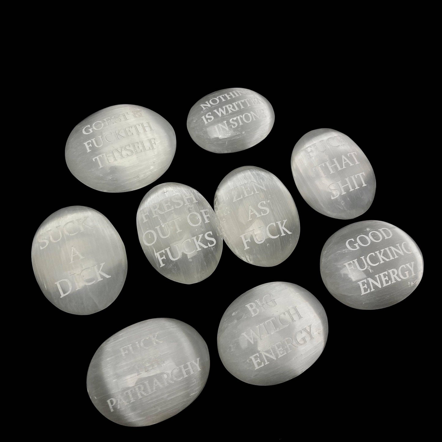 Nine smooth, polished white stone pendants with engraved humorous and feminist quotes, arranged on a solid black background.