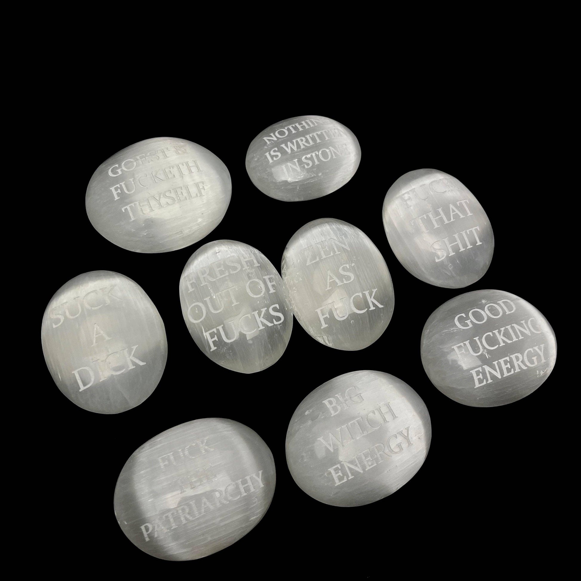 Nine smooth, polished white stone pendants with engraved humorous and feminist quotes, arranged on a solid black background.