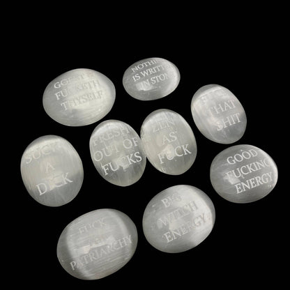 Nine smooth, polished white stone pendants with engraved humorous and feminist quotes, arranged on a solid black background.