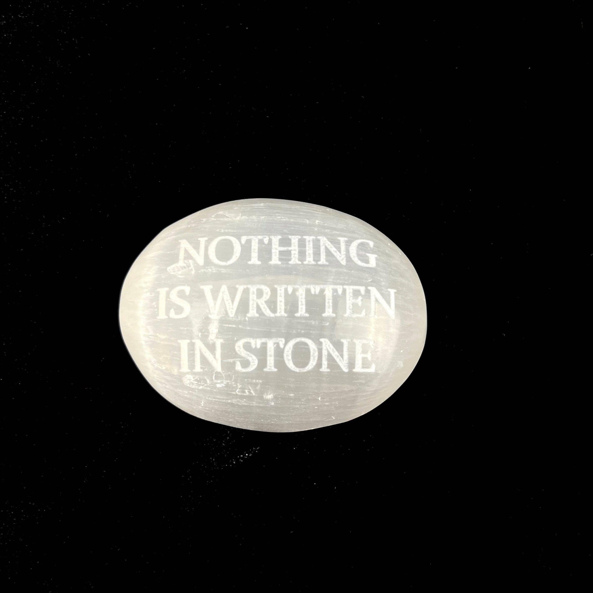 A smooth, oval-shaped translucent white stone with the phrase 'NOTHING IS WRITTEN IN STONE' engraved in white lettering, set against a solid black background.