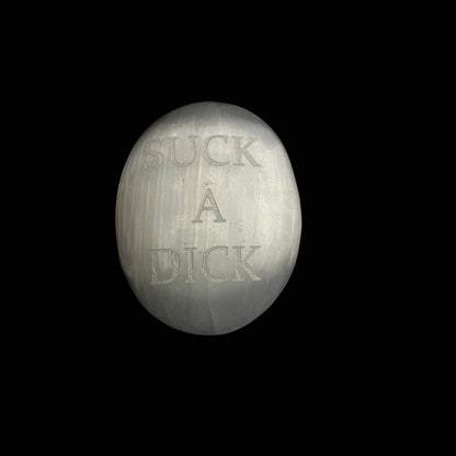 A metallic silver oval object with the words 'SUCK A DICK' embossed on it, centered against a solid black background.