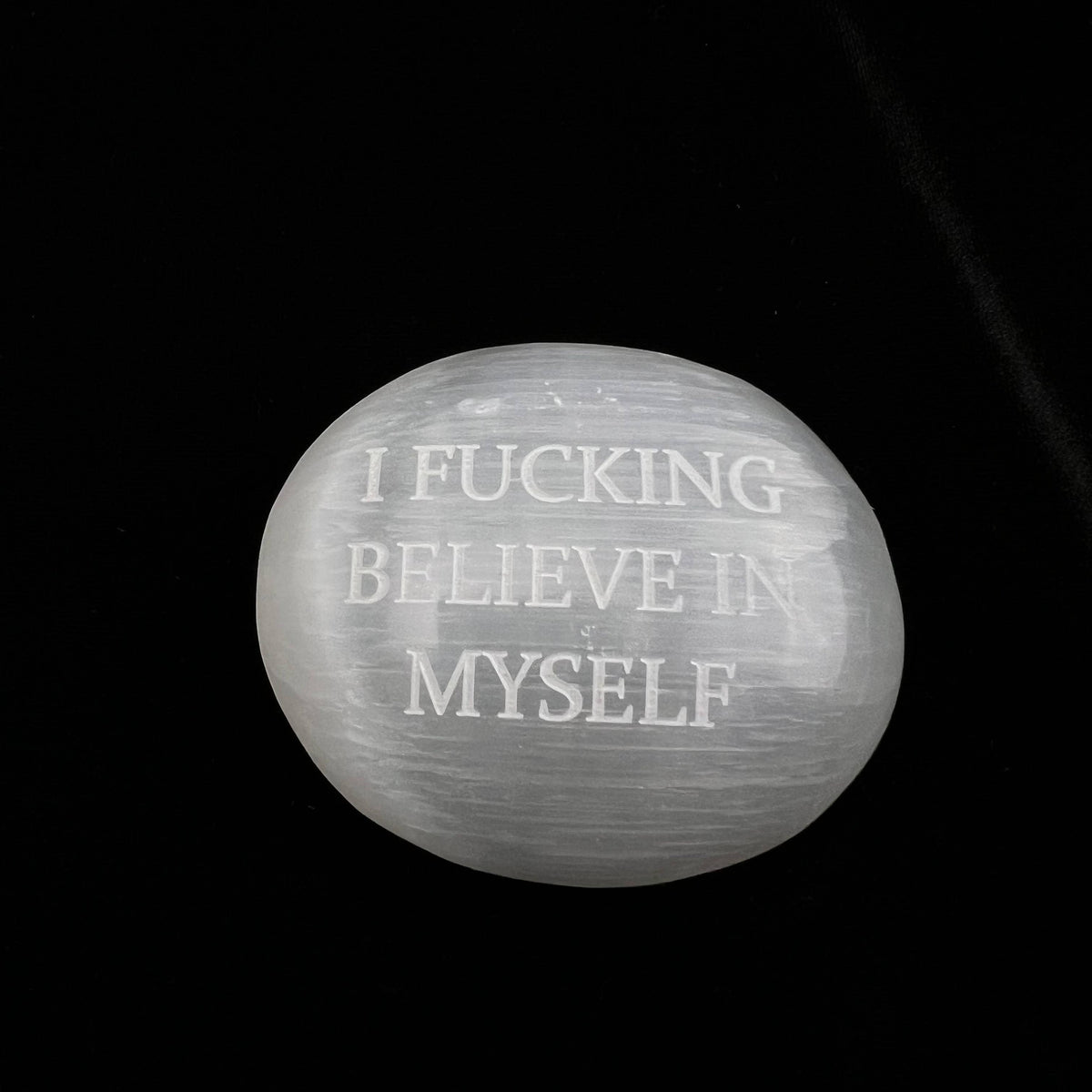 A white selenite stone with the text 'I FUCKING BELIEVE IN MYSELF' engraved on it, set against a solid black background.