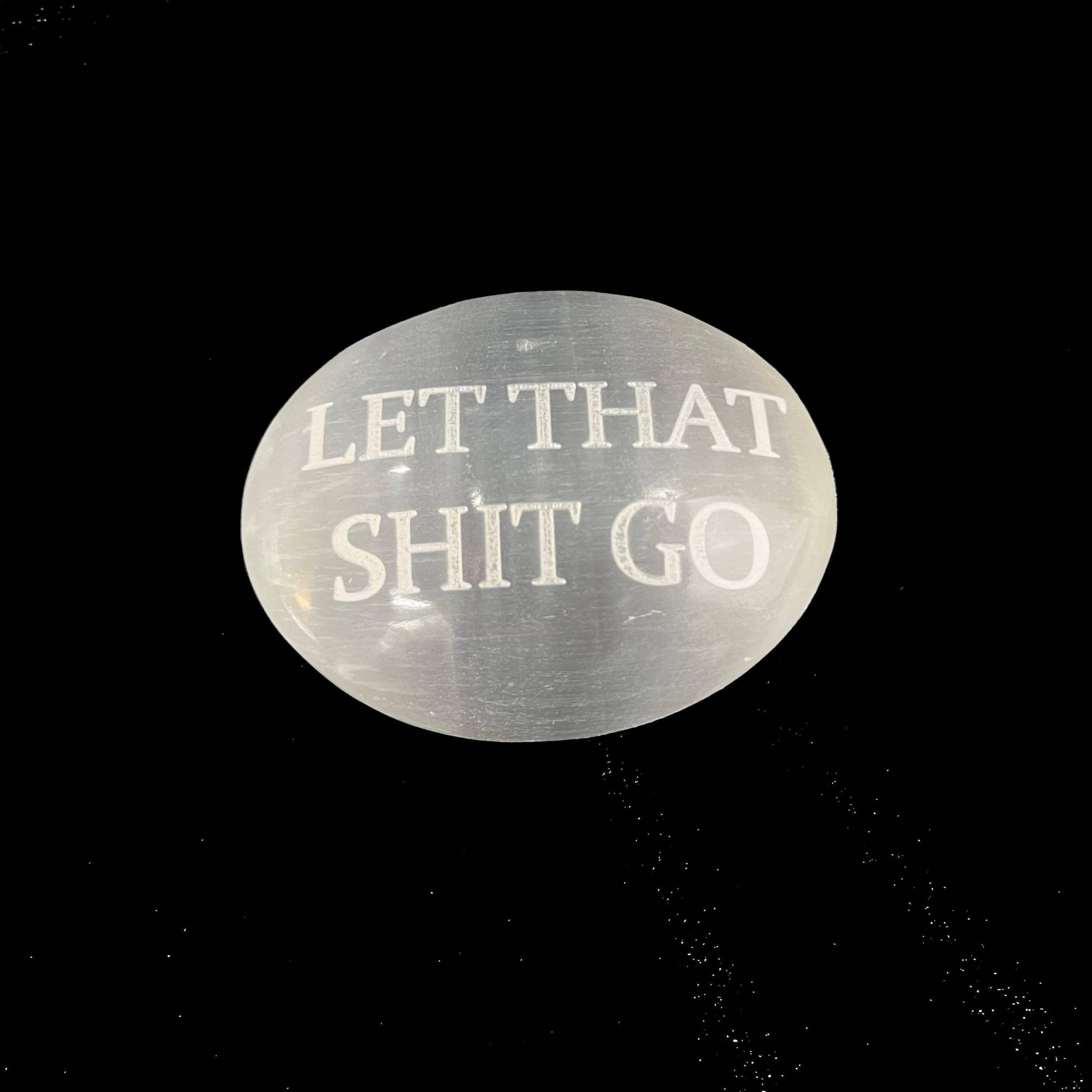 A polished oval white stone with the phrase 'LET THAT SHIT GO' engraved in white lettering, set against a solid black background.