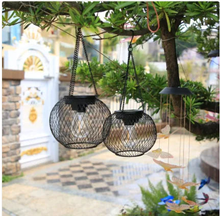 Two hanging solar lights on a tree branch with a garden and building in the background.
