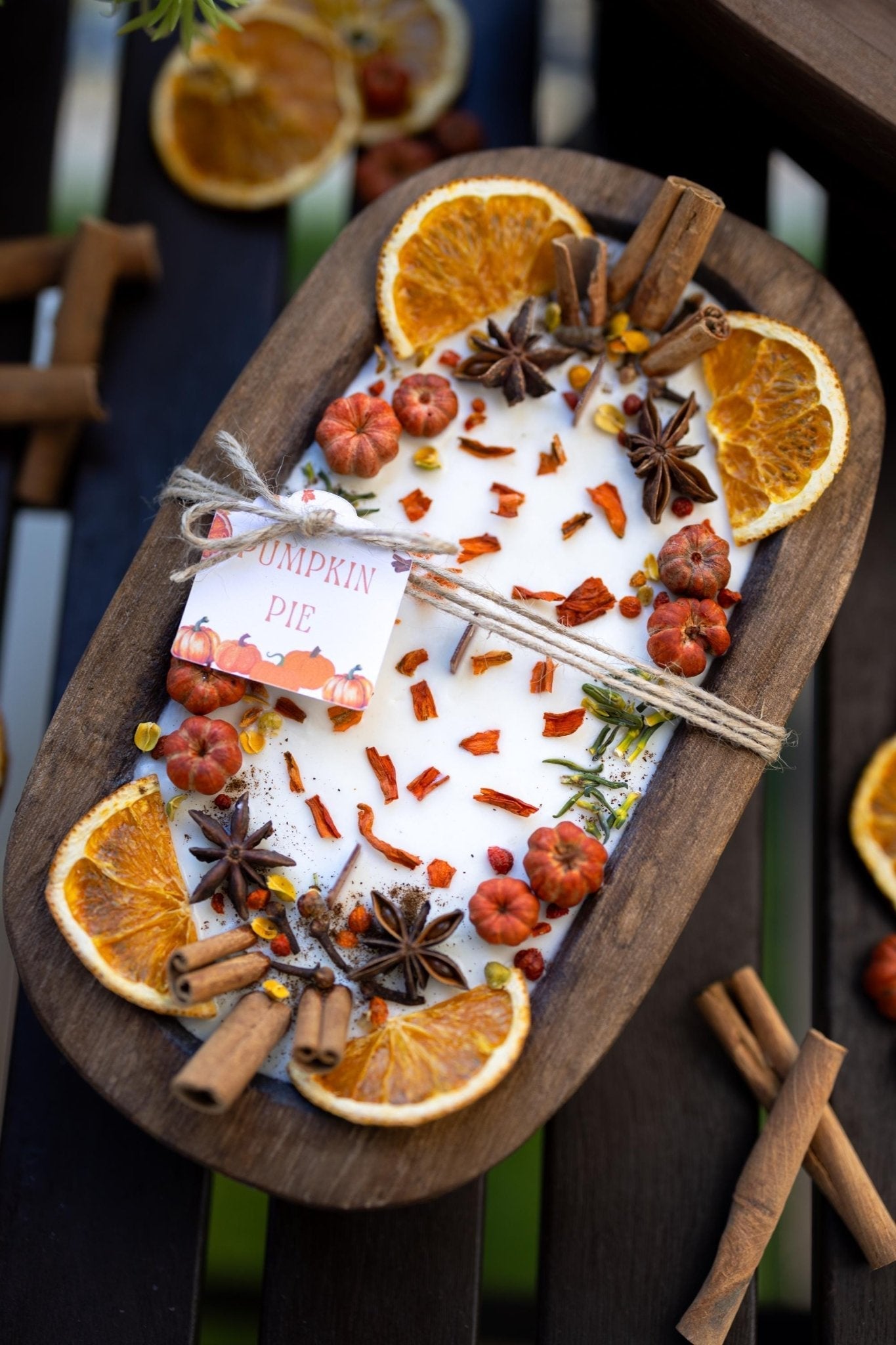 An oval wooden tray holding a white candle topped with dried orange slices, cinnamon sticks, star anise, small red berries, and scattered orange peel pieces. A small tag reads 'Pumpkin Pie.