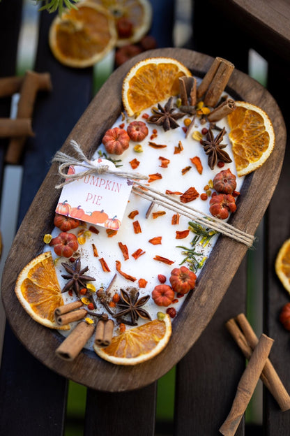 An oval wooden tray holding a white candle topped with dried orange slices, cinnamon sticks, star anise, small red berries, and scattered orange peel pieces. A small tag reads 'Pumpkin Pie.