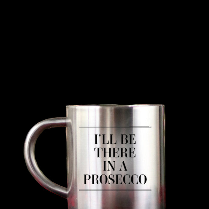 Silver mug with 'I'll be there in a Prosecco' text on a red checkered tablecloth with a Christmas tree background.