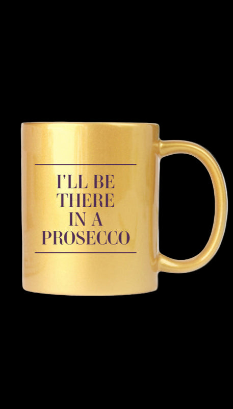 Gold mug with 'I'll be there in a prosecco' text on a white surface with a plant in the background