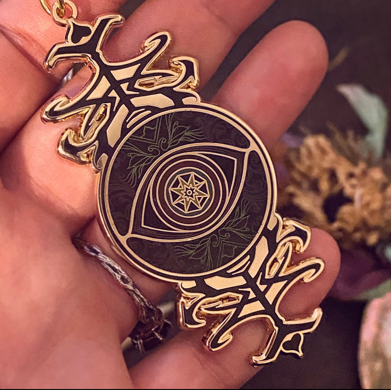 A close-up of a hand holding a circular pendant with an eye design and ornate gold-toned side elements. The pendant has a dark background with green leaf patterns and a star motif in the center.