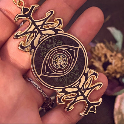 A close-up of a hand holding a circular pendant with an eye design and ornate gold-toned side elements. The pendant has a dark background with green leaf patterns and a star motif in the center.