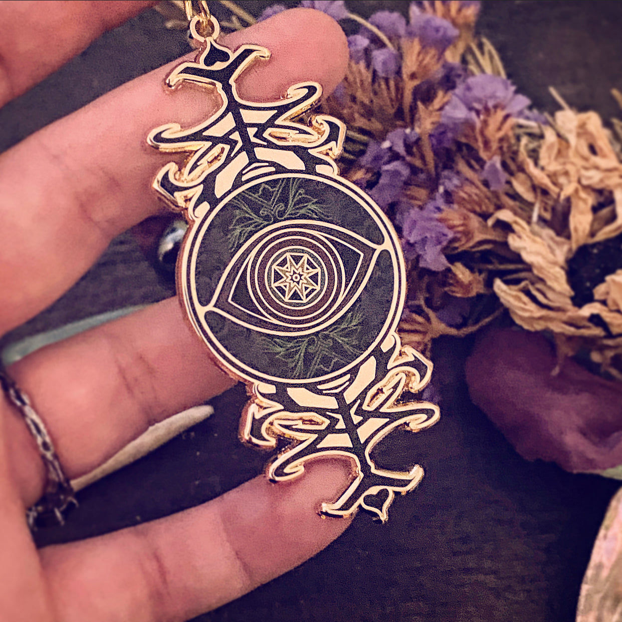 A gold-toned pendant featuring a central eye design with a star and crescent moon motif, held in a hand against a dark background with dried flowers.